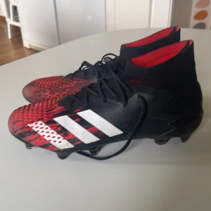 Adidas fotbollsskor Predator - Säljer ett par Adidas Predator fotbollsskor i svart och rött med vita detaljer. Skorna har snörning, hög krage och klassiska tre ränder på sidan. Perfekta för dig som vill ha bra grepp på planen och snygg design.