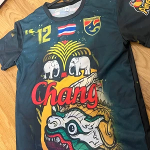 Thailand fotbollströja Chang #12 - Unik thailändsk fotbollströja med tryck av elefanter, drake och Chang-logga. Tröjan har nummer 12, Thailand-flagga och gult mönster. Märke på ärmen och stor text på ryggen. Perfekt för dig som gillar färgstarka och detaljerade motiv.