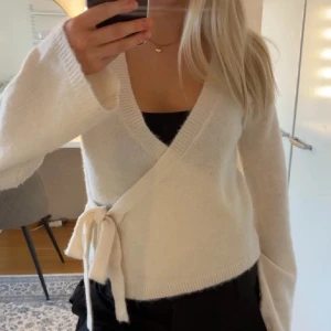 Vit omlottstickad kofta med knyt - Säljer en supermjuk vit stickad kofta med omlottmodell och knyt i sidan. Tröjan har v-ringning och långa ärmar, perfekt att styla över ett linne eller topp. Enkel och stilren design som passar till mycket.