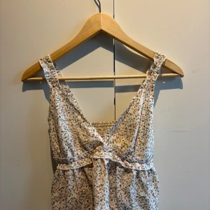 Blommig ärmlös linnetopp - Supersöt ärmlös linnetopp med blommigt mönster i pastellfärger. Toppen har breda axelband, v-ringning och en liten rosett framtill. Perfekt för varma dagar och ger en romantisk känsla till outfiten.