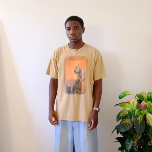 Beige t-shirt med tryck 'Children of Uganda' - En vintage t-shirt med tryck av en man som spelar trummor. T-shirten är i en beige färg och har en rund halsringning. Trycket är i en brun och orange färg och visar en man som spelar trummor med en klubba. T-shirten är i gott skick och har inga synliga defekter.  Vintage T-shirt   Children of Uganda  Size M/L  Length:73cm Chest: 51cm Arm-Length: 22cm