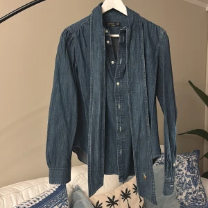 Jeansskjorta med rosett från Polo Ralph Lauren - Säljer en snygg jeansskjorta från Polo Ralph Lauren med knappar framtill och stor knytrosett vid kragen. Skjortan har långa ärmar och klassisk broderad logga nertill. Perfekt för dig som gillar denim och vill ha något extra.