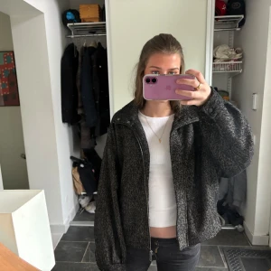 Mörkgrå kappa med knappar - Säljer en mörkgrå kappa med diskret fiskbensmönster. Jackan har hög krage, långa ärmar och knäppning framtill. Passformen är oversized och materialet känns som ullblandning. Perfekt för kyligare dagar.