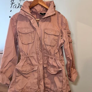 Rosa parkajacka med dragkedja från H&M - Säljer en rosa parkajacka från H&M med dragkedja och fyra stora fickor framtill. Jackan har lång ärm, hög krage och justerbar midja med dragsko. Perfekt för vår och höst.