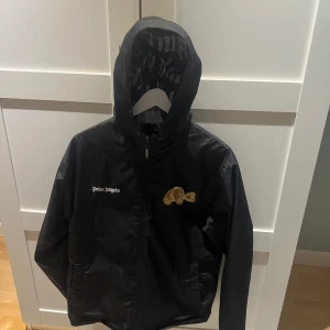 Svart vindjacka från Palm Angels - Säljer en svart vindjacka från Palm Angels med huva och dragkedja. Jackan har ett broderat nallemärke på bröstet och Palm Angels-logga på vänster sida. Insidan är fodrad med stort Palm Angels-tryck.