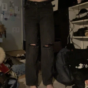 Svarta jeans med hål på knäna - Säljer ett par svarta jeans med raka ben och slitna hål på båda knäna. Jeansen har hög midja och klassisk femficksdesign. Perfekta för en avslappnad och trendig stil.