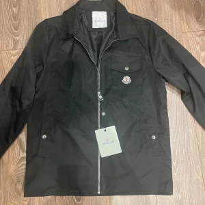 Svart jacka från Moncler med dragkedja - Snygg svart jacka från Moncler med klassisk krage och silverfärgad dragkedja framtill. Jackan har en bröstficka med knapp och Moncler-logga, samt två sidofickor med knappar. Perfekt för en stilren look.