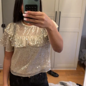 Glittrig topp med volang från Lindex  - Säljer en glittrig topp i ljusbeige med paljetter och volangdetaljer över bröstet och axlarna. Toppen har korta ärmar och rund halsringning. Perfekt för dig som vill sticka ut lite extra!