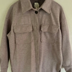Ljuslila overshirt med fickor - Säljer en ljuslila overshirt med två stora bröstfickor och klassisk krage. Jackan har lång ärm och stängs med en knapp framtill. Perfekt för lager-på-lager och enkel att styla till olika outfits.