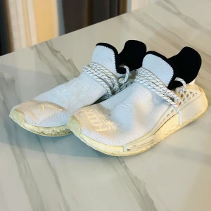 Adidas Pharrell Williams Human Race sneakers vita - Säljer ett par vita Adidas Pharrell Williams Human Race sneakers med svart häl och broderad text på ovandelen. Skorna har snörning och en platt sula med rutmönster undertill. Perfekta för dig som gillar unika sneakers med cool design.