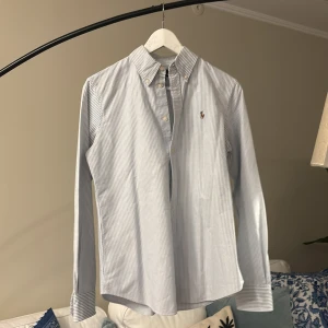 Blåvit randig skjorta från Polo Ralph Lauren - Klassisk blåvit randig skjorta från Polo Ralph Lauren med broderad logga på bröstet. Skjortan har lång ärm, knappar framtill och button-down krage. Tillverkad i bomull och har en normal passform.