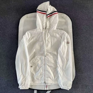 Vit vindjacka från Moncler  - Snygg vit vindjacka från Moncler med huva och dragkedja. Jackan har Moncler-logga på ärmen och röd-blå rand på huvan. Perfekt för dig som vill ha en stilren och sportig look. Fickor med dragkedja och elastiska muddar vid ärmslut.
