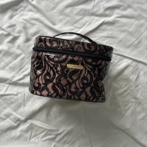 Necessär från Victoria's Secret med spetsmönster - Säljer en snygg necessär från Victoria's Secret med svart spetsmönster över rosa botten. Väskan har dragkedja med guldfärgad detalj och ett praktiskt handtag upptill. Perfekt för smink eller småprylar.