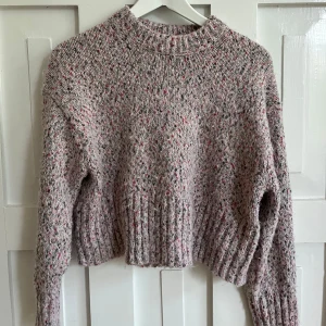 Melerad croppad stickad tröja från H&M - Säljer en croppad stickad tröja från H&M i melerad beige med inslag av rosa, rött och grönt. Tröjan har lång ärm, ribbad kant vid ärmslut och nederkant samt en högre rund halsringning.