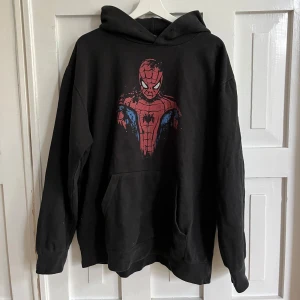 Svart hoodie med Spiderman-tryck - Svart hoodie med stor Spiderman-illustration på bröstet i rött, blått och vitt. Tröjan har huva och en stor magficka framtill.