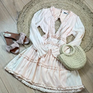 Ljusrosa omlottklänning med volangdetaljer - Supersöt ljusrosa klänning med lång ärm, volangdetaljer och v-ringning framtill. Klänningen har öppen rygg och fina spetskanter, samt en kort, svängande kjol. Perfekt för sommarens alla tillfällen!  Är i M men passar även S