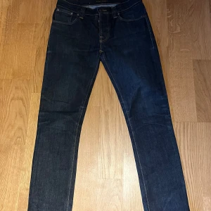 Mörkblå jeans Grim Tim från Nudie Jeans - Säljer ett par mörkblå Grim Tim jeans från Nudie Jeans i modellen Org. Dry Navy. Klassisk femficksdesign, raka ben och orangea sömmar. Tillverkade av 99% ekologisk bomull och 1% elastan för lite stretch.