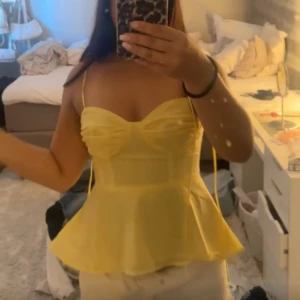 Gul croppad linnetopp med volang - Snygg gul linnetopp med smala axelband och volang nertill. Toppen har en söt, figurnära passform och lätt v-ringad byst. Perfekt för varma dagar och somriga outfits.