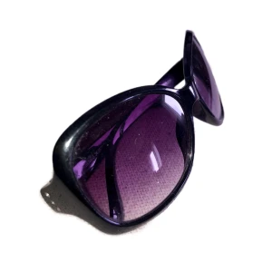 Svarta solglasögon med lila tonade glas - Stilrena solglasögon med svart båge och stora, rundade glas i lila ton. Bågarna har en glansig finish och diskreta rhinestones vid sidorna. Perfekta för att ge en cool touch till din look.