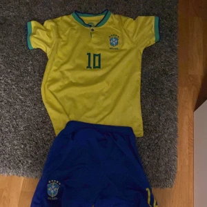 Gul Brasilien fotbollströja med blå shorts - Säljer en klassisk gul Brasilien fotbollströja med blå detaljer och nummer 10 på bröstet. Tröjan har korta ärmar och blå krage. Tillhörande blå shorts med Brasilien-logga och nummer 10. Perfekt för fotbollsälskare!