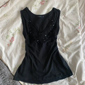 Svart linne med spetsdetaljer från Vila - Säljer ett svart linne från Vila med v-ringning och dekorativ spets framtill, prydd med paljetter. Toppen är ärmlös och har en figurnära passform, perfekt att styla till olika outfits.