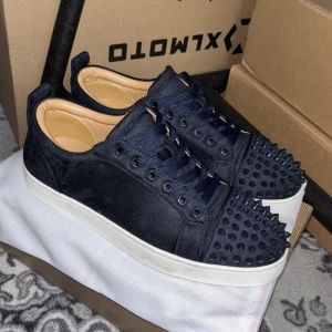 Mörkblåa sneakers med nitar från Christian Louboutin - Säljer ett par mörkblåa sneakers från Christian Louboutin med matta nitar på tån och ovandelen. Skorna har klassisk röd sula, snörning och är tillverkade i mocka. Perfekta för dig som vill sticka ut med en edgy stil.