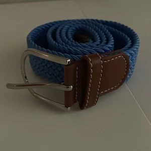 Flätat blått bälte med bruna skinn-detaljer - Snyggt flätat bälte i blått med bruna skinn-detaljer och silverfärgat spänne. Perfekt accessoar för att lyfta din outfit med en färgklick. Passar till jeans eller chinos.