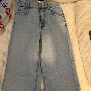 Ljusblå jeans med råa kanter - Säljer ett par ljusblå jeans med raka, vida ben och fransiga kanter nertill. Jeansen har klassisk femficksdesign och knappgylf. Perfekta för en avslappnad och trendig look. Dock är de lite slitna längst ner då de var för långa för mig