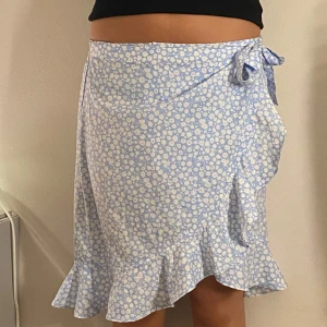 Ljusblå blommig omlottkjol med volang - Supersöt ljusblå omlottkjol med vitt blommönster och volangkant. Kjolen knyts i sidan med en rosett och har en luftig och somrig känsla. Perfekt för dig som gillar romantisk stil och vill ha något lätt och bekvämt.