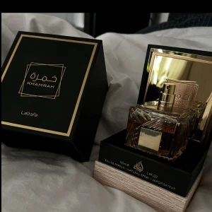 Khamrah Eau de Parfum från Lattafa - Exklusiv parfym i elegant fyrkantig glasflaska med gulddetaljer och matchande svart och guldig förpackning. Doften är intensiv och lyxig, perfekt för dig som vill sticka ut. Flaskan rymmer 100 ml och har en stilren design med spegelinslag.