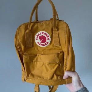 Gul Kånken Mini ryggsäck från Fjällräven - Säljer en ikonisk gul Kånken Mini ryggsäck från Fjällräven. Väskan har klassisk fyrkantig form, bärhandtag upptill, justerbara axelremmar och en rund röd logotyp framtill. Praktisk framficka med dragkedja och slitstarkt material.