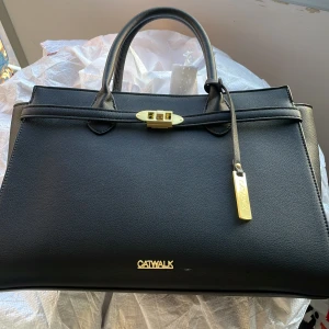 Svart handväska från Catwalk med gulddetaljer - Stilren svart handväska från Catwalk med guldfärgade detaljer och lås framtill. Väskan har dubbla handtag, dragkedjeficka på baksidan och rymligt innerfack. Pris kan diskuteras i DM. Skriv gärna till mig innan du vill köpa!