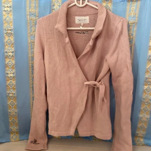 Ljusrosa omlottkofta från Capri Collection - Säljer en ljusrosa stickad omlottkofta från Capri Collection. Koftan har lång ärm, knytband i midjan och fina detaljer med broderad text och stjärna på ärmen och ryggen. Perfekt för lager-på-lager och en mjuk känsla.