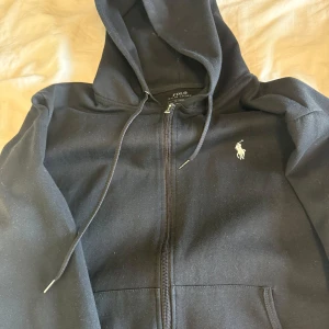 Svart zip hoodie från Polo Ralph Lauren - Svart hoodie från Polo Ralph Lauren med dragkedja och klassisk broderad logga på bröstet. Tröjan har huva med snörning och fickor framtill. Perfekt för en avslappnad stil. 