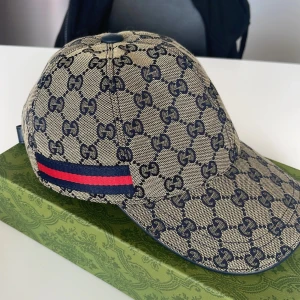 Beige keps med monogram från Gucci - Snygg beige keps från Gucci med klassiskt GG-monogram i mörkblått och röd-blå rand på sidan. Justerbar rem baktill och insida i brunt tyg. Perfekt för dig som vill ha en exklusiv accessoar.