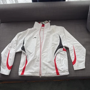 Vit vindjacka med röda och svarta detaljer - Säljer en vit vindjacka med röda och svarta grafiska detaljer längs sidorna och ärmarna. Jackan har dragkedja framtill, hög krage och två fickor. Perfekt för träning eller som en sportig vardagsjacka.