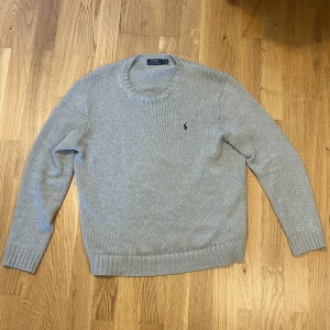 Ljusgrå stickad tröja från Polo Ralph Lauren - Säljer en klassisk ljusgrå stickad tröja från Polo Ralph Lauren med rund halsringning och den ikoniska broderade loggan på bröstet. Tröjan har långa ärmar och ribbade muddar vid ärmslut och nederkant. Perfekt för lager-på-lager-stil.