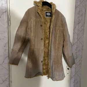 Beige pälsfodrad skinnjacka från Livelo - Säljer en beige skinnjacka från Livelo med mjukt, ljust pälsfoder och knappar framtill. Jackan har längre modell och markerade sömmar som ger en cool vintagekänsla. Perfekt för dig som gillar klassisk stil med twist.