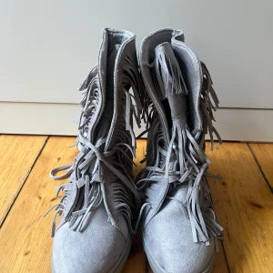 Grå boots med fransar från Siader - Säljer ett par grå boots från Siader med snygga fransdetaljer och snörning framtill. Skorna har en platt sula och är tillverkade i mocka. Perfekta för dig som vill sticka ut med din stil!