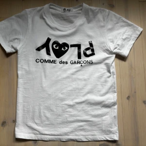 Vit t-shirt från Comme des Garçons Play - Vit t-shirt från Comme des Garçons Play med svart tryck på bröstet där det står 'PLAY' och 'COMME des GARÇONS' samt ett hjärta med ögon. Klassisk rund hals och korta ärmar. Perfekt för dig som vill ha en riktigt snygg t-shirt till sommaren priset är inte hugget i sten