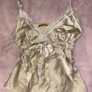 Champagnefärgad spetslinnetopp från Eden - Säljer en champagnefärgad linnetopp från Eden med smala axelband, spetsdetaljer längs v-ringningen och en söt rosett framtill. Toppen är gjord i glansigt material, passar S/XS