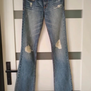 Blå bootcut jeans från Abercrombie - Säljer ett par blå bootcut jeans från Abercrombie med slitna detaljer på knäna och klassisk femficksdesign. Jeansen har låg midja och är tillverkade i denim med snygg tvätt och slitningar framtill. Midjemåttet 38cm, innerbenslängd 81cm