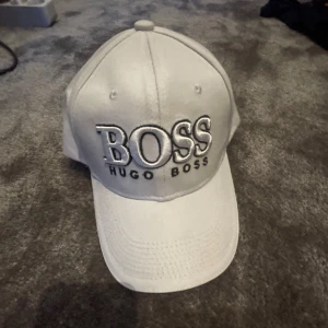 Vit keps från Hugo Boss - Snygg vit keps från Hugo Boss med stor broderad BOSS-logga framtill. Justerbar rem baktill för perfekt passform. Skick 9/10 aldrig använd bara lite skrap märke som ni ser på sista bilden. Klassisk böjd skärm och ventilerande hål upptill. Perfekt accessoar för en stilren look. Kan sänkas vid snabb affär.