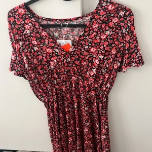 Blommig röd och svart klänning med v-ringning - Supersöt klänning med röd och rosa blommigt mönster på svart botten. V-ringad med korta ärmar och smock i midjan för en smickrande passform. Perfekt för sommarens alla tillfällen!