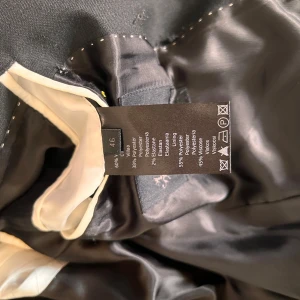 Svart kavaj från Riley - Snygg svart kavaj från Riley i storlek M. Kavajen har två knappar framtill, smala slag och flera fickor. Tillverkad i en blandning av ull och polyester med ett slätt foder. Perfekt för dig som vill ha en klassisk och stilren look.