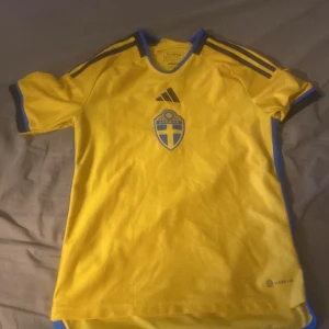 Svenska landslagets fotbollströja från Adidas - Gul kortärmad fotbollströja (Sverige) säsongen 23/24 från Adidas med blå detaljer och svenska landslagets emblem på bröstet. Klassisk design med tre blå ränder på axlarna och blå kant vid ärmslut och krage. Perfekt för dig som vill visa ditt stöd för Sverige. Köpt för 550kr