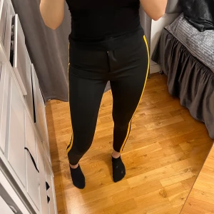 Svarta tighta byxor med gula revärer från Bershka - Ett par svarta tighta byxor från Bershka med två gula revärer längs sidorna. Byxorna har dragkedja och knapp framtill och är i stretchigt material. 