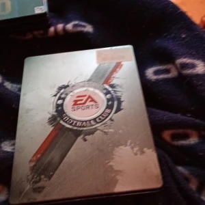 EA Sports Football Club Steelbook - Säljer en exklusiv EA Sports Football Club steelbook med snyggt tryck i silver, rött och blått. Perfekt för samlare av fotbollsspel eller dig som gillar unika spelboxar. Metallmaterial med robust känsla.