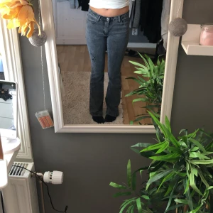 Grå/svarta bootcut jeans med slitning - Säljer ett par grå bootcut jeans med medel midja och en snygg slitning på ena benet. Jeansen har klassiska fem fickor och stängs med dragkedja och knapp. Perfekta för dig som gillar en lite retro vibe.