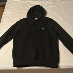 Svart hoodie från Lacoste - Säljer en svart hoodie från Lacoste med klassisk krokodillogga på bröstet. Tröjan har huva med dragsko och en stor magficka. Perfekt för dig som gillar stilrena och bekväma plagg.